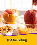 Apples with caramel coating on sticks, one dipped in caramel, with a bowl of caramel and snacks in the background.