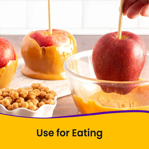 Apples with caramel coating on sticks, one dipped in caramel, with a bowl of caramel and snacks in the background.