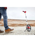 Built Different 12 Inch Digital Measuring Wheel featuring smooth rubber tread, spoked frame, and a worker's boot on rough terrain.
