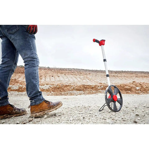 Built Different 12 Inch Digital Measuring Wheel featuring smooth rubber tread, spoked frame, and a worker's boot on rough terrain.