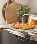 Front view: 12-inch aluminum pizza peel with handle lifting a cheese pizza into a countertop oven.