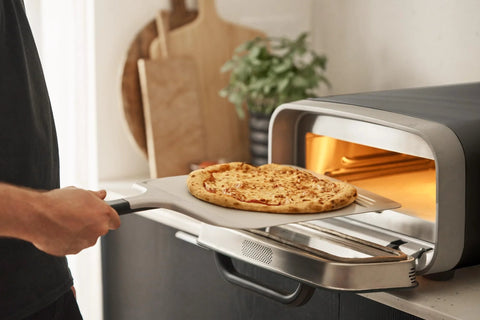 Front view: 12-inch aluminum pizza peel with handle lifting a cheese pizza into a countertop oven.