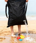 Man holding a black bag with water pouring out onto sand with toys, depicting 2-In-1 Sand Free Beach Blanket And Tent, 79 By 83 Inch Waterproof Picnic Blanket with Built-In Inflatable Pillows, Quick Inflation No Pump, 360 Degree Sandproof Walls, Fits 6 To 10 Seated Or 4 To 6 Reclined For Family Camping Travel Backyard.