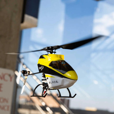 120S2 RC Helicopter BNF with SAFE Technology flying outdoors, yellow and black design, carbon fiber frame visible, clear sky background