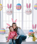 9 Piece Easter Paper Lantern Set With Bunny And Egg Designs, Reusable 10 Inch Pastel Hanging Lanterns For Home Garden Or Outdoor Easter Party, Kid Friendly Decor And Party Favors For Spring Fun displayed in a window setting with people wearing bunny ears.