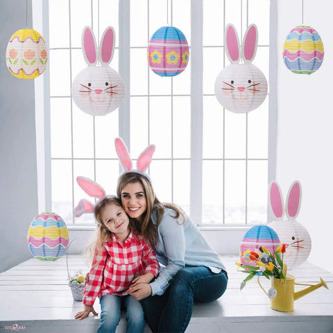 9 Piece Easter Paper Lantern Set With Bunny And Egg Designs, Reusable 10 Inch Pastel Hanging Lanterns For Home Garden Or Outdoor Easter Party, Kid Friendly Decor And Party Favors For Spring Fun displayed in a window setting with people wearing bunny ears.