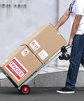 Aluminum folding hand truck with red wheels carrying two cardboard boxes labeled FRAGILE.