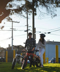 Two electric bikes with riders in a park; white and red bikes with helmets.