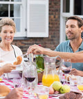 Family enjoying a meal at an outdoor table with drinks and fruit on a red checked tablecloth.