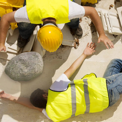 Worker assisting another lying down on construction site wearing yellow hard hats and reflective vests.