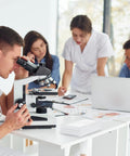 Laboratory scene with people using microscopes and laptops around a table, engaged in scientific research and discussion.