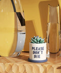 Small ceramic planter with text 'PLEASE DON'T DIE' holding a succulent, placed on a wood shelf beside glass vases.