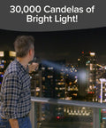 Man uses Monster Bright Rechargeable Spotlight With 10 Modes And Turbo Boost To 100 Percent Built In Kickstand And USB Phone Charger Ideal For Camping And Emergencies 9 Inch Black Flashlight As Seen On TV to illuminate city at night, showcasing its 30,000 candelas of brightness.