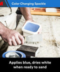 Person holding putty knife and container of color-changing spackling compound from High Strength Drywall Hole Repair Kit With Color Changing Spackling Compound 12 Oz Includes Putty Knife Patch And Sanding Pad Fast Drying For Interior And Exterior Use Easy Fix For Small Holes on a workbench.