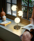 Three Light Brass Bedside Lamp With Mid Century Globe Shade In White And Gold illuminating a cozy reading corner by a window.