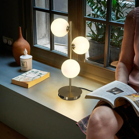 Three Light Brass Bedside Lamp With Mid Century Globe Shade In White And Gold illuminating a cozy reading corner by a window.