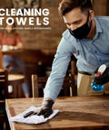 Man cleaning a wooden table with white towel from Absorbent Quick Dry Bar Mop Towels Pack Of 60, 14x17 Commercial Grade Cotton Cleaning Rags For Kitchen And Bar, White Reusable Shop Towels For Heavy Duty Cleaning And Spill Cleanup, wearing gloves and mask in a restaurant setting.