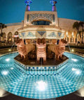 Illuminated fountain with lion statues and patterned tiles in front of ornate building at night.