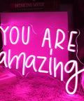 Pink You Are Amazing LED Neon Light Sign For Wall Decor In Bedrooms Living Rooms Bars Parties Classrooms USB Powered 16.9 By 10.6 Inches Hanging Or Table Display glowing on a fuzzy surface.