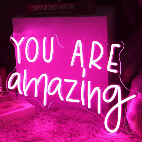 Pink You Are Amazing LED Neon Light Sign For Wall Decor In Bedrooms Living Rooms Bars Parties Classrooms USB Powered 16.9 By 10.6 Inches Hanging Or Table Display glowing on a fuzzy surface.