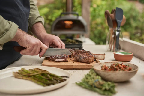 Person grilling with Karu 2 Pro Multi Fuel Pizza Oven For Backyard Grilling Wood Or Gas Fired 16 Inch Stone Pizza Maker in background