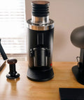 Heavy Duty 64Mm Flat Burr Electric Coffee Grinder Designed For Single Dosing Habits With Anti Popcorn Guard And Zero Static Hiccups grinding on a wooden table.