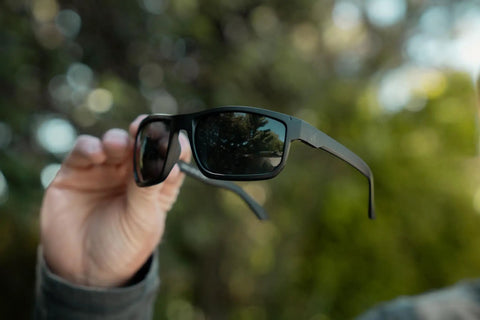 Kickass Z87 Certified Wayfarer Safety Sunglasses Featuring Hella Effective Antifog Smoke Lenses And Totally Shatterproof Matte Black Frames Built Tough For Male Workers held in hand outdoors.