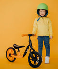 Child standing beside orange balance bike with Kid Bike Seat Replacement Cushion for 12 and 14 Inch Balance Bikes visible, black.