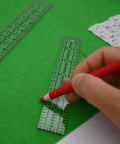 Hand using a red pencil on a green felt background with Dead Simple Acrylic Quilting Rulers And Nifty Fabric Cutting Templates For Patchwork Projects.