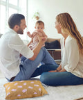 Parents playing with baby on floor with patterned pillow nearby in a cozy, light-filled living room.