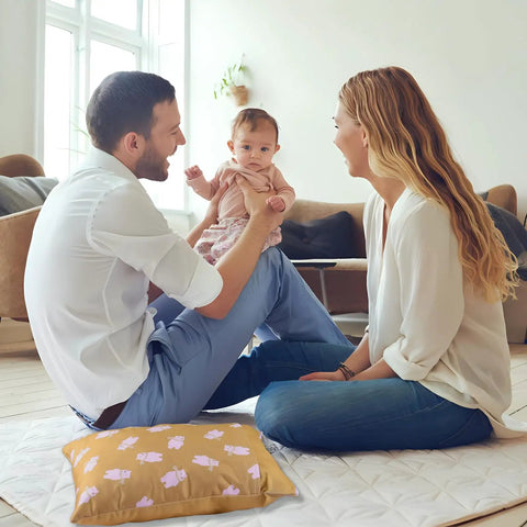 Parents playing with baby on floor with patterned pillow nearby in a cozy, light-filled living room.