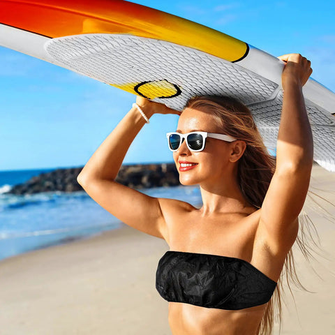Woman holding surfboard on beach wearing black strapless top, smiling in sunglasses, sunny blue sky, ocean waves in background.