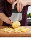 Black-handled potato cutter with stainless steel comb blades slicing a potato into fries on a wooden cutting board