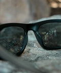 Kickass Z87 Certified Wayfarer Safety Sunglasses Featuring Hella Effective Antifog Smoke Lenses and Totally Shatterproof Matte Black Frames Built Tough For Male Workers resting on a rock.