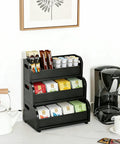 Black bamboo three-layer stackable tea bags organizer on kitchen counter next to coffee maker, holding assorted tea bags.