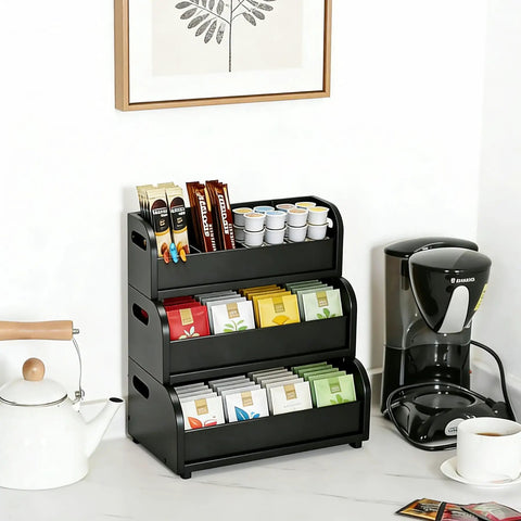 Black bamboo three-layer stackable tea bags organizer on kitchen counter next to coffee maker, holding assorted tea bags.