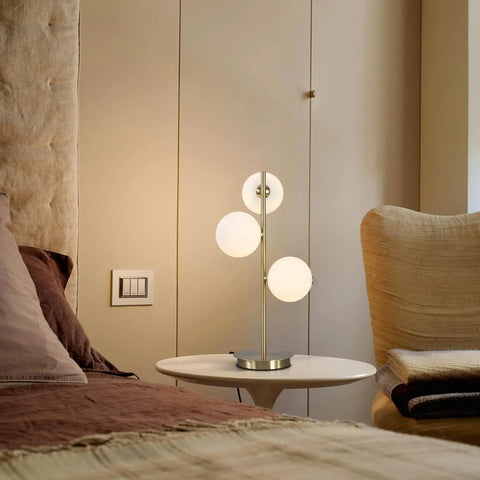 Three Light Brass Bedside Lamp With Mid Century Globe Shade In White And Gold For Reading In Office Living Room Dresser And Bathroom G9 Base Perfect Retro Modern Vibe And Cozy Nightstand Ambience shown on a bedside table next to a beige armchair and bed.