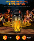 Flame Lamp Speaker With Multicolor Lights For Outdoors Patio Pool Camping Waterproof Torch Lantern 7 Lighting Modes And Music Sync RGB Glow, placed on table, with group enjoying a campfire vibe in the background.