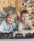 Two boys playing with Electric Train Set For Kids With Steam Locomotive And Cargo Cars Tracks And Railway Kit under Christmas tree