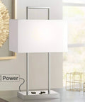Jaymes Chrome Table Lamp With White Fabric Shade, 26-Inch Tall on desk, showing power outlet feature