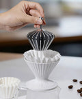 Hand placing reusable stainless steel filter into white pour over coffee dripper cup. Solid 3 In 1 Metal Pour Over Coffee Dripper Gift Set With Reusable Stainless Steel Filter And 25 Basket Papers For Hassle Free Brewing Smooth Daily Grind Or Chill Travel Vibes At Home And On The Road.