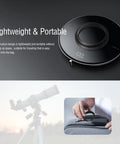 Round portable wireless charger on surface, telescope in background and hand inserting charger into bag showing its compact design.