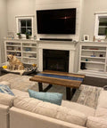 A cozy living room with a beige couch, wooden coffee table, and a large TV above a white fireplace. A dog sits near the fireplace.
