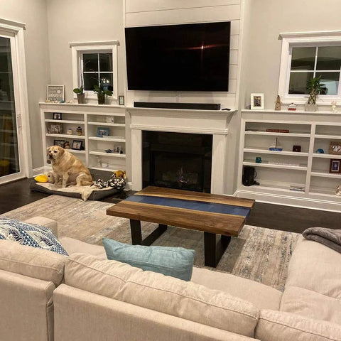 A cozy living room with a beige couch, wooden coffee table, and a large TV above a white fireplace. A dog sits near the fireplace.
