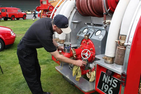 Person polishing fire truck equipment outdoors with metal cleaner, showing hoses and gauges, on grass field.