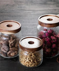 Keep Your Stash Fresh With These Top Notch Sealed Tea Canisters And Coffee Bean Jars Crafted From Durable Glass Super Simple Kitchen Pantry Storage Essentials That Are Actually Worth The Buy Hella Convenient: Three glass canisters with wooden lids, storing dried flowers and herbs.