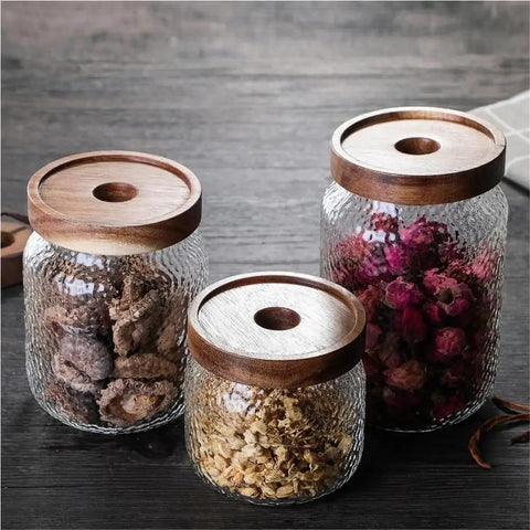 Keep Your Stash Fresh With These Top Notch Sealed Tea Canisters And Coffee Bean Jars Crafted From Durable Glass Super Simple Kitchen Pantry Storage Essentials That Are Actually Worth The Buy Hella Convenient: Three glass canisters with wooden lids, storing dried flowers and herbs.