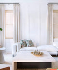 Modern living room with white sofa, light wood coffee table, tall plants, and large windows with curtains