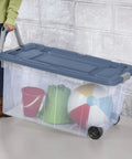 Person carrying a clear wheeled storage tote filled with toys, featuring a secure lid and easy grip handles.