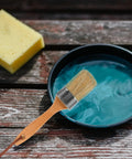 Two Pack Wood Handle Paint Brush Set With Synthetic Bristles Oval Stain Brush For Deck And Fence Smooth Wall Painting Easy Clean Up Great For Furniture And Latex Paint Applications, shown with a brush in turquoise paint, sponge nearby on a wooden surface.